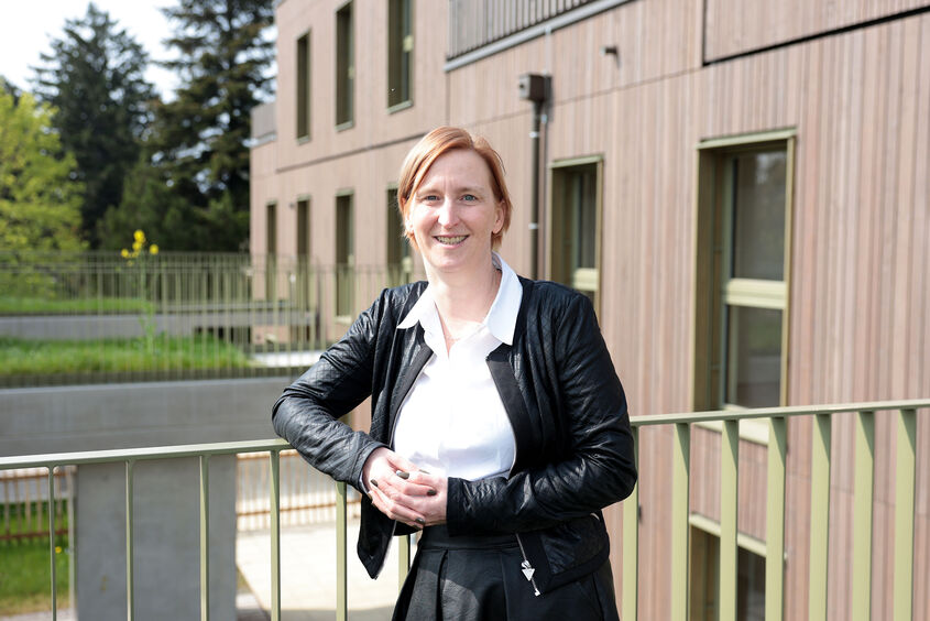 Frau mit schwarzer Lederjacke und weißem Hemd lehnt an einem Geländer vor einem modernen Wohngebäude mit Holzfassade