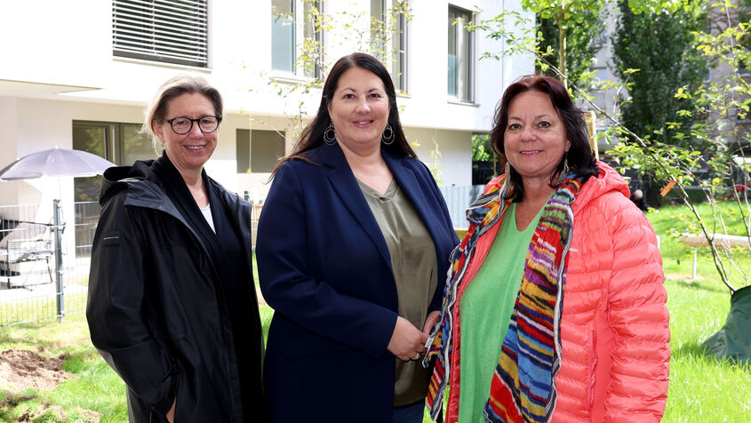Wiener Wohnen Direktorin Karin Ramser, Vizebürgermeisterin und Wohnbaustadträtin Kathrin Gaál und wohnpartner-Bereichsleiterin Claudia Huemer stehen im begrünten Innenhof des neuen Gemeindebaus. Die drei sind trotz des offensichtlich freundlichen Wetters herbstlich gekleidet und lächeln freundlich in die Kamera.