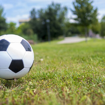 Schwarz-weißer Fußball auf einem Rasen mit unscharfem Hintergrund und einer Person in Shorts und Turnschuhen
