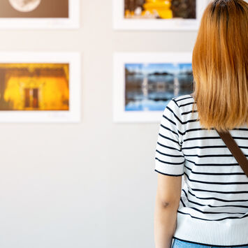 Person von hinten in gestreiftem Shirt und mit Umhängetasche betrachtet gerahmte Fotos an einer weißen Galeriewand.