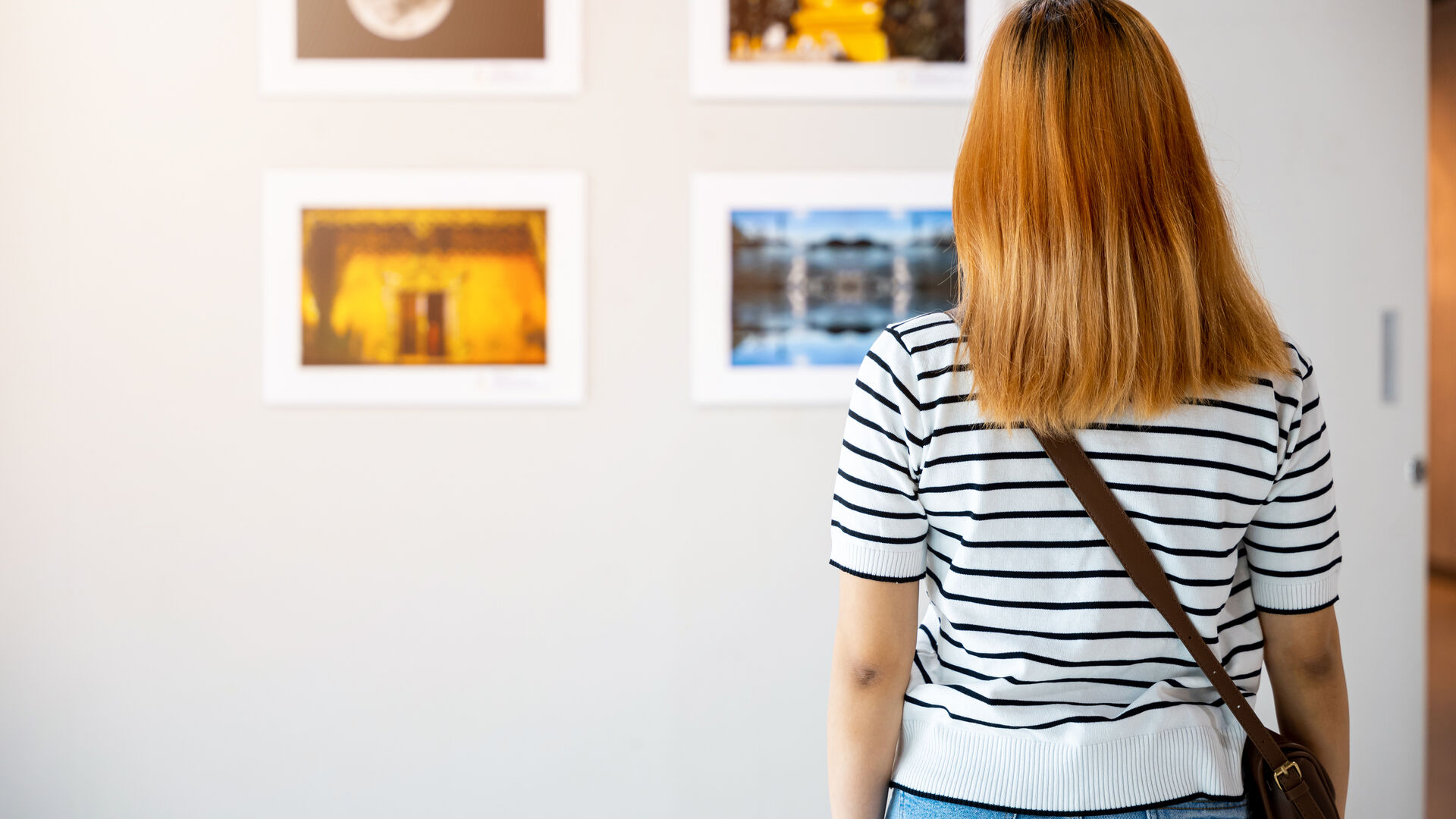 Person von hinten in gestreiftem Shirt und mit Umhängetasche betrachtet gerahmte Fotos an einer weißen Galeriewand.