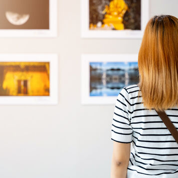 Person von hinten in gestreiftem Shirt und mit Umhängetasche betrachtet gerahmte Fotos an einer weißen Galeriewand.