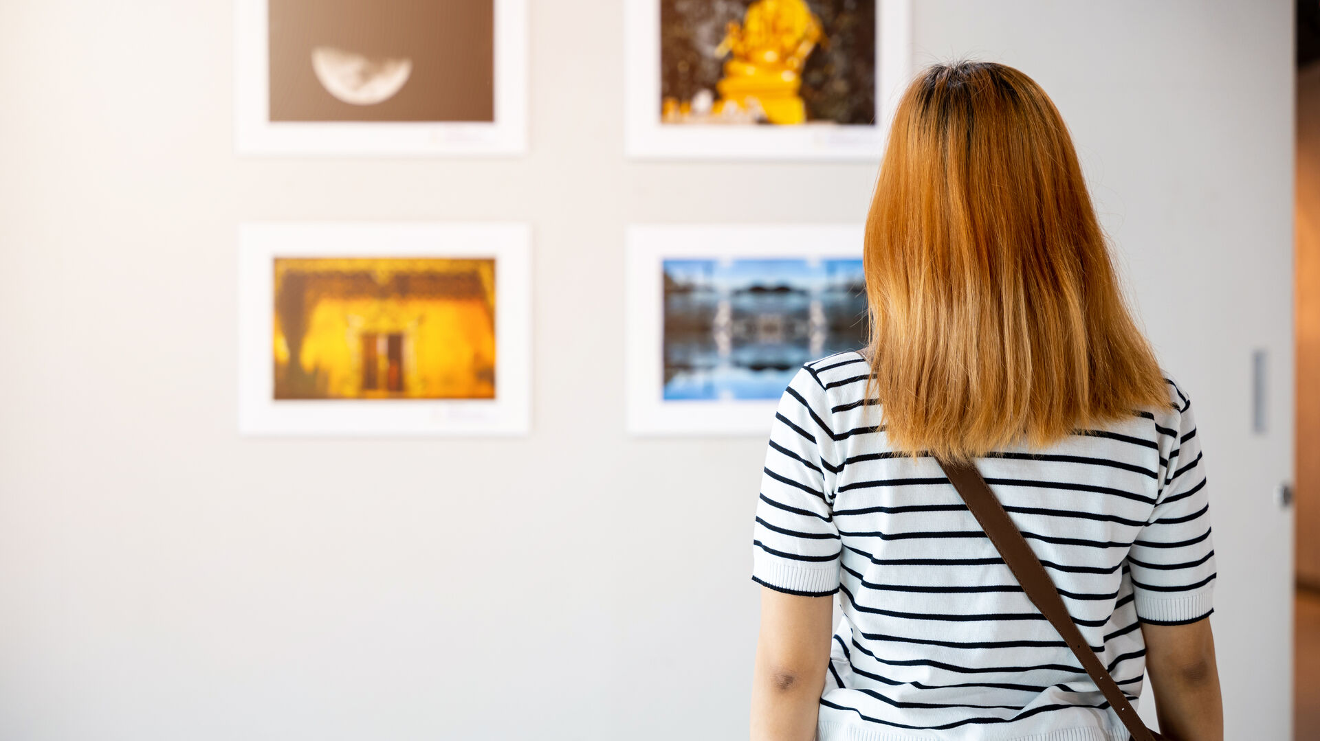 Person von hinten in gestreiftem Shirt und mit Umhängetasche betrachtet gerahmte Fotos an einer weißen Galeriewand.