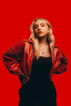Eine Person mit langem Haar steht in einem schwarzen Kleid und mit roter Lederjacke vor rotem Hintergrund. Sie schaut in die Kamera und hat die Arme in die Hüften gestreckt.