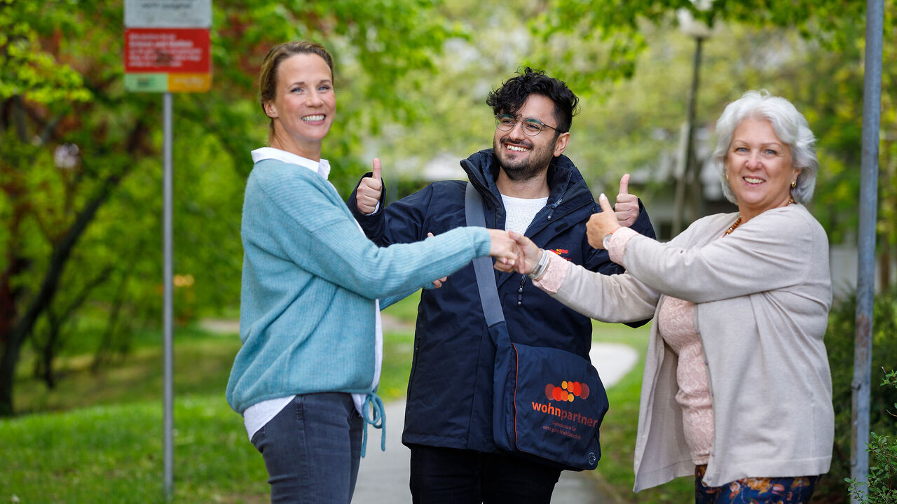 Drei Personen stehen auf einem asphaltierten Weg im Grünen. Die beiden Frauen stehen einander gegenüber seitlich zur Kamera, reichen einander die Hand und lächeln in die Kamera. Hinter den Frauen, in der Mitte des Bildes steht ein wohnpartner-Mitarbeiter mit wohnpartner-Jacke und -Tasche, lächelt ebenfalls in die Kamera und hält beide Daumen hoch. Die ältere der beiden Frauen hält ebenfalls den Daumen hoch. Das Imagebild soll die erfolgreiche Mediation zwischen zwei Parteien durch einen Mitarbeiter von wohnpartner vermitteln.
