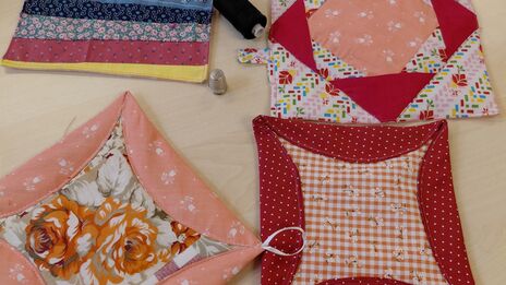 Vier bunte, quadratische Patchwork-Topflappen auf einem hellen Tisch mit einer Schere, einem schwarzen Garnknäuel und einem Fingerhut