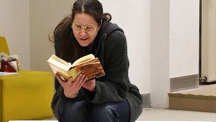 Eine Person sitzt auf einer Bank und liest in einem Buch. Sie trägt eine runde goldene Brille