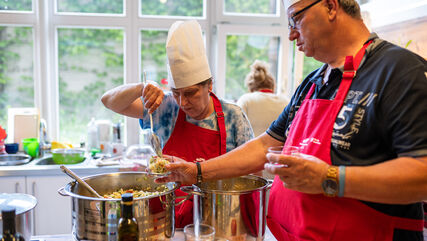 2 Bewohner*innen beim Abfüllen von Essen in kleine Glasschüsselchen, sie tragen rote Kochschürzen und weiße Kochhüte
