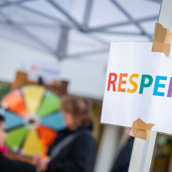 Ein Zettel mit der Aufschrift "RESPEKT", dahinter sind Kinder zu sehen, die an einem Glücksrad drehen.