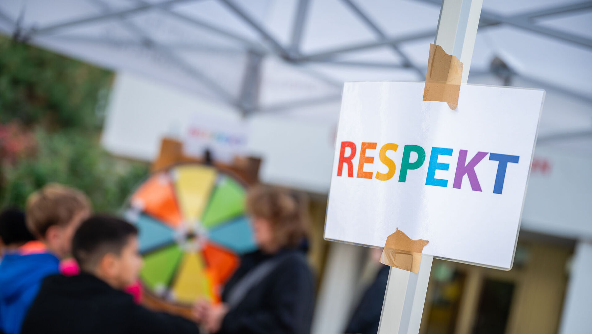 Ein Zettel mit der Aufschrift "RESPEKT", dahinter sind Kinder zu sehen, die an einem Glücksrad drehen.