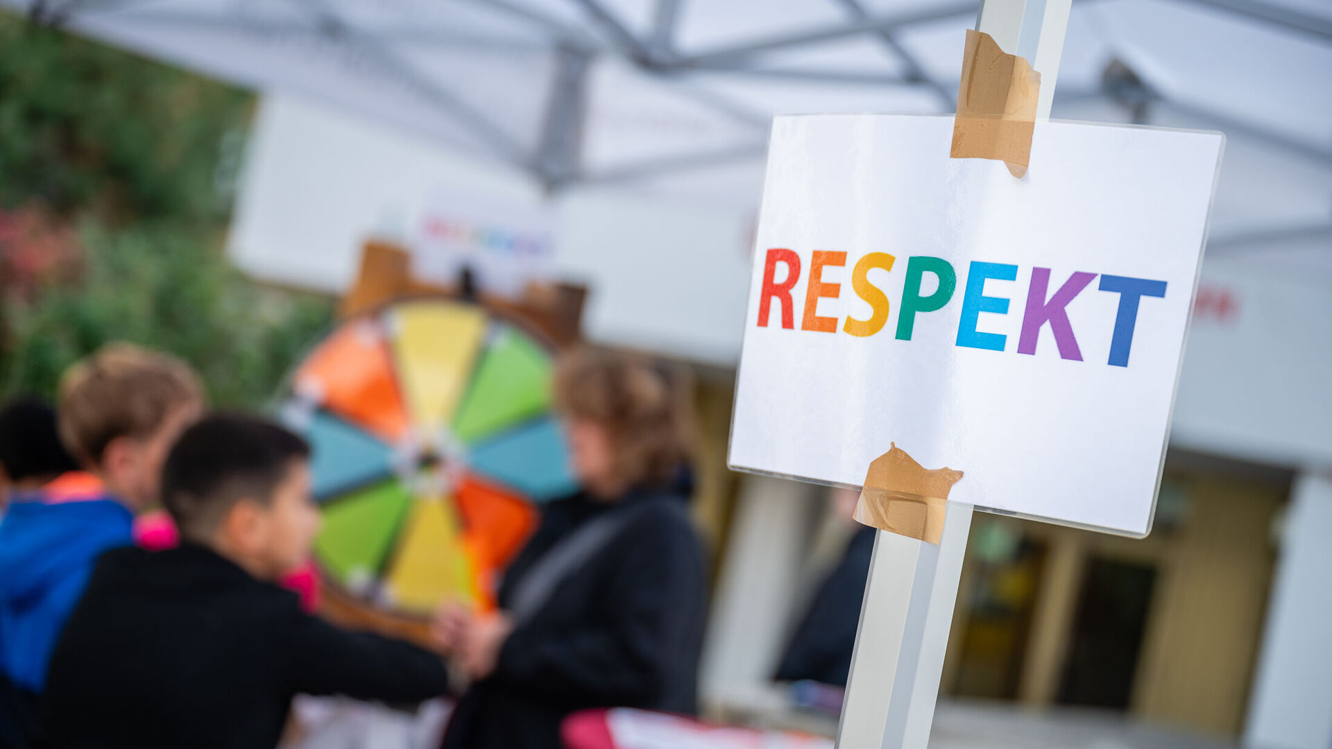 Ein Zettel mit der Aufschrift "RESPEKT", dahinter sind Kinder zu sehen, die an einem Glücksrad drehen.