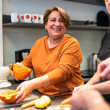 Kochgruppe in einer wohnpartner-Küche, im Zentrum eine ältere lachende Dame, die Kürbis schneidet.