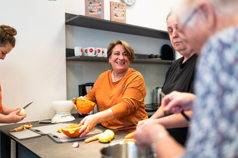 Kochgruppe in einer wohnpartner-Küche, im Zentrum eine ältere lachende Dame, die Kürbis schneidet.