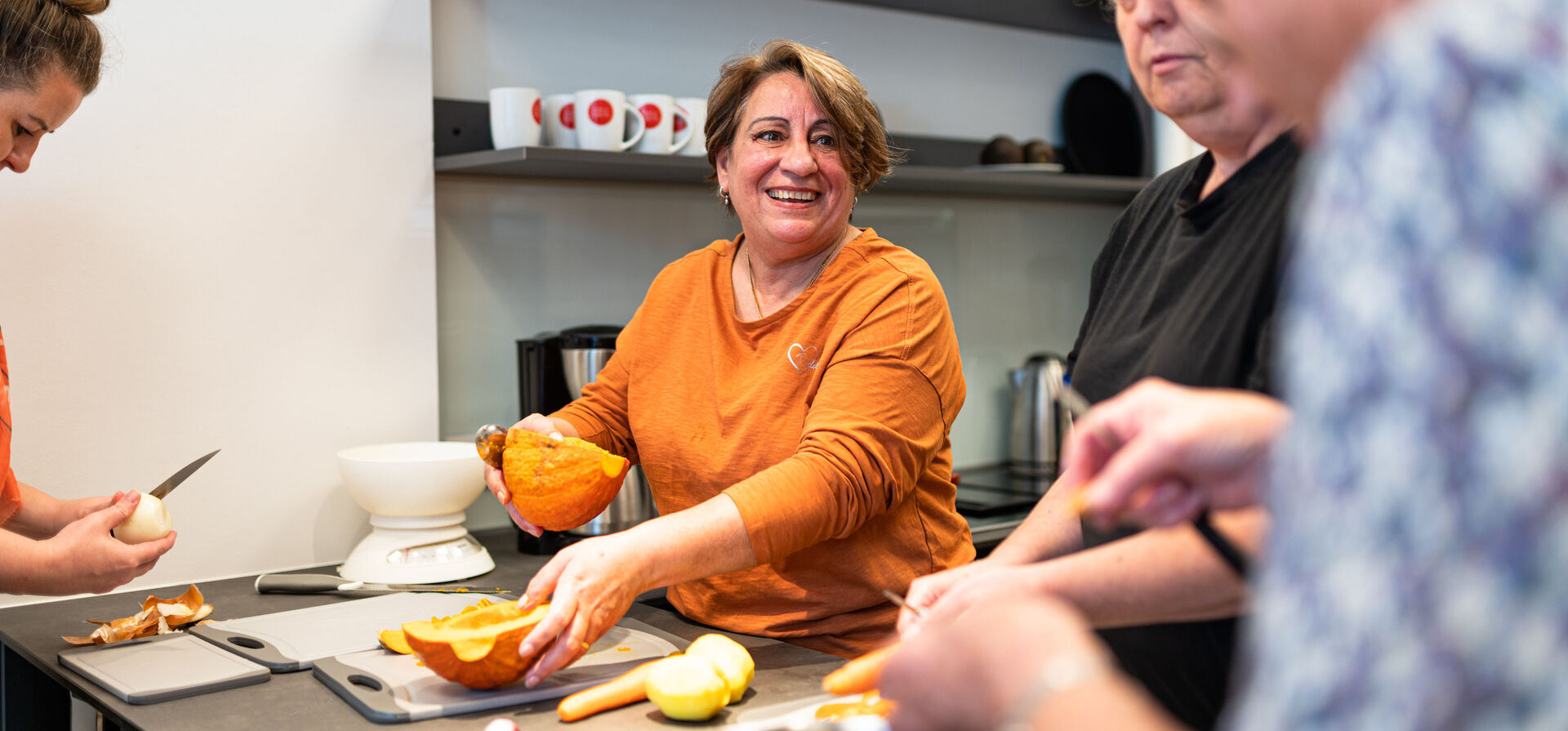 Kochgruppe in einer wohnpartner-Küche, im Zentrum eine ältere lachende Dame, die Kürbis schneidet.