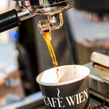 Nahaufnahme einer Siebträgermaschine: Barista schenkt Kaffee in einen Pappbecher mit der Aufschrift "Café Wien" ein.