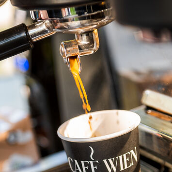 Nahaufnahme einer Siebträgermaschine: Barista schenkt Kaffee in einen Pappbecher mit der Aufschrift "Café Wien" ein.