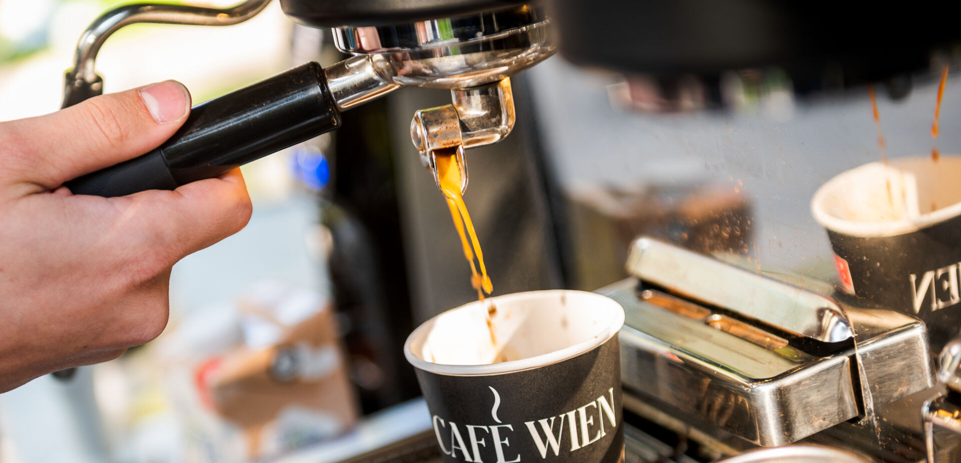 Nahaufnahme einer Siebträgermaschine: Barista schenkt Kaffee in einen Pappbecher mit der Aufschrift "Café Wien" ein.