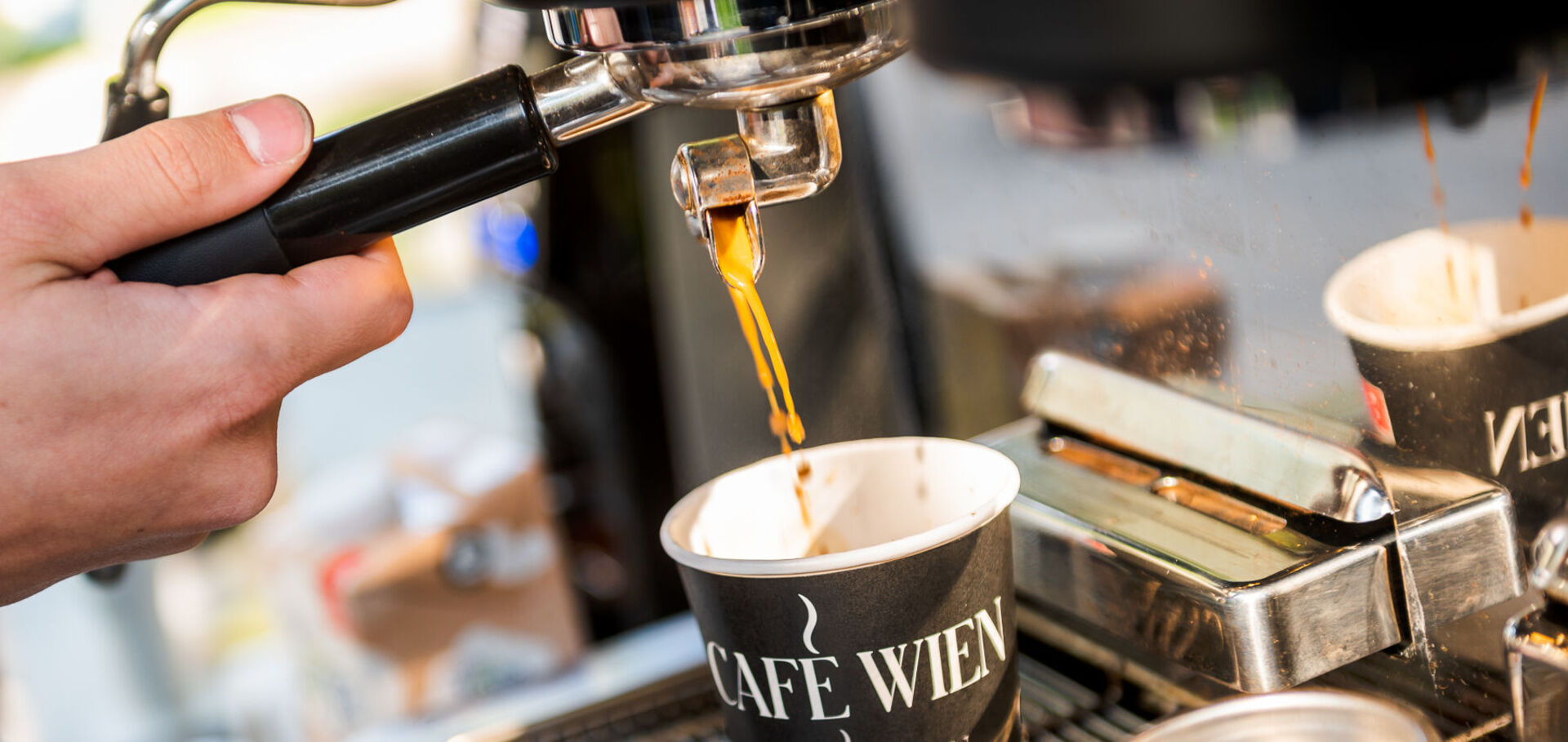 Nahaufnahme einer Siebträgermaschine: Barista schenkt Kaffee in einen Pappbecher mit der Aufschrift "Café Wien" ein.