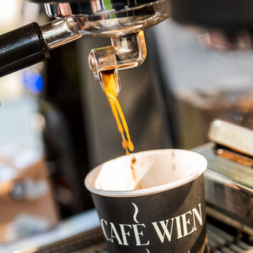 Nahaufnahme einer Siebträgermaschine: Barista schenkt Kaffee in einen Pappbecher mit der Aufschrift "Café Wien" ein.