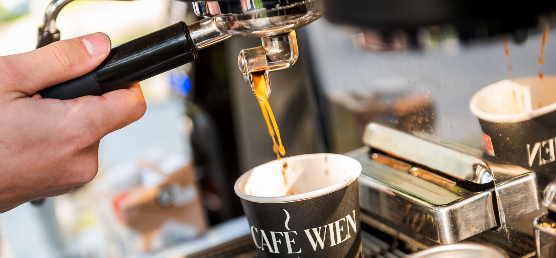 Nahaufnahme einer Siebträgermaschine: Barista schenkt Kaffee in einen Pappbecher mit der Aufschrift "Café Wien" ein.