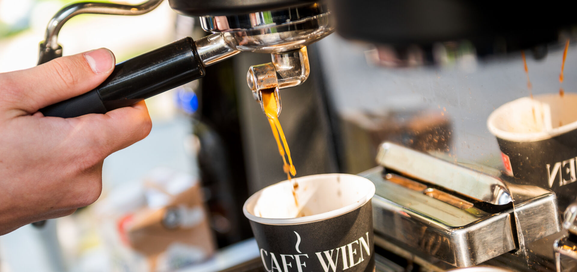 Nahaufnahme einer Siebträgermaschine: Barista schenkt Kaffee in einen Pappbecher mit der Aufschrift "Café Wien" ein.