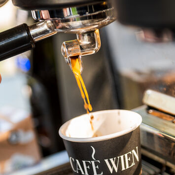 Nahaufnahme einer Siebträgermaschine: Barista schenkt Kaffee in einen Pappbecher mit der Aufschrift "Café Wien" ein.