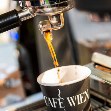 Nahaufnahme einer Siebträgermaschine: Barista schenkt Kaffee in einen Pappbecher mit der Aufschrift "Café Wien" ein.