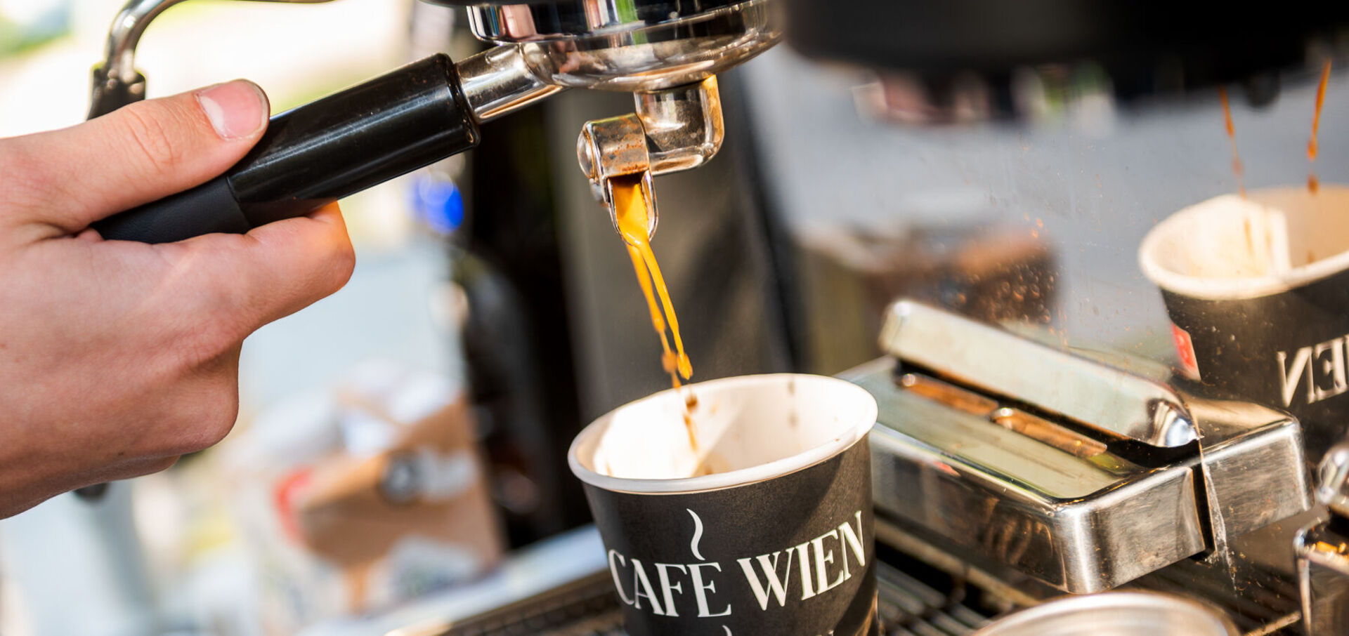 Nahaufnahme einer Siebträgermaschine: Barista schenkt Kaffee in einen Pappbecher mit der Aufschrift "Café Wien" ein.