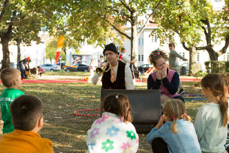 Zwei verkleidete Schauspieler (ein Mann, eine Frau) im Park spielen vor einer Gruppe von Kindern Trompete und Mundharmonika