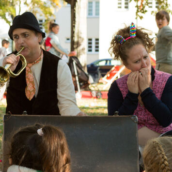 Zwei verkleidete Schauspieler (ein Mann, eine Frau) im Park spielen vor einer Gruppe von Kindern Trompete und Mundharmonika