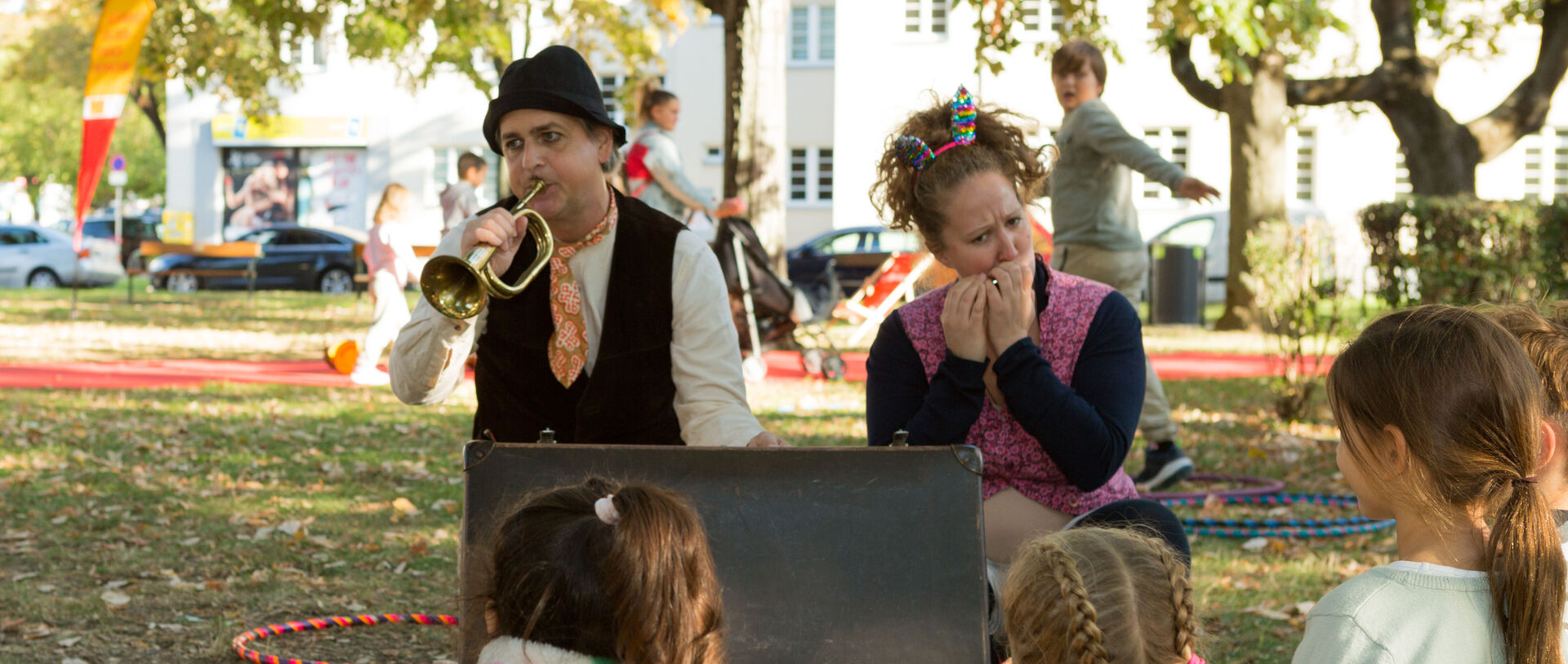 Zwei verkleidete Schauspieler (ein Mann, eine Frau) im Park spielen vor einer Gruppe von Kindern Trompete und Mundharmonika