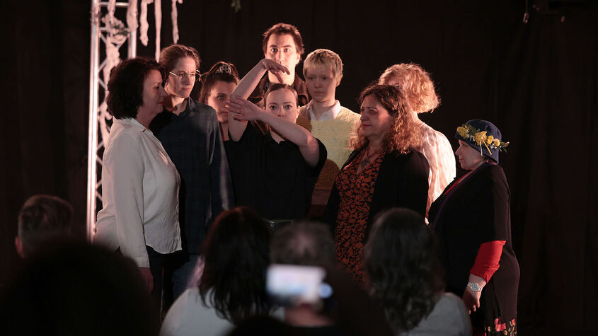 Mehrere Personen stehen knapp beisammen und blicken auf eine in der Mitte platzierte Person die mit ihren Armen die Äste eines Baumes darstellt. Der Hintergrund ist schwarz, im Vordergrund erkennt man Personen sitzend von hinten, die auf die Personengruppe schauen. 