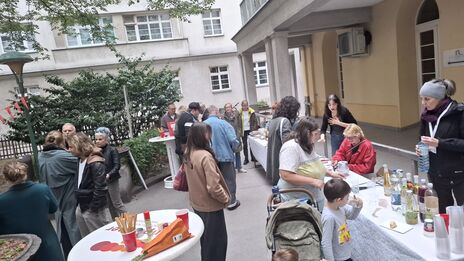 Menschen stehen an Tischen mit Getränken und Snacks in einem Innenhof mit Gebäuden im Hintergrund