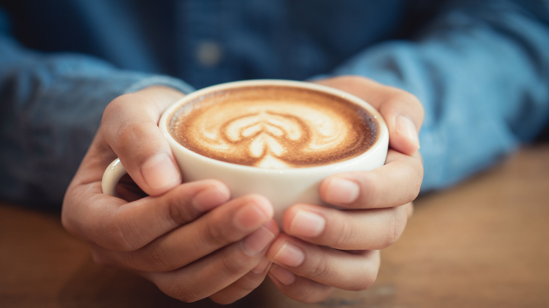 Zwei Hände halten eine Tasse Kaffee mit Milchschaum und Herzmuster