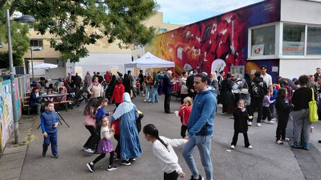 Menschen verschiedener Altersgruppen, darunter Kinder und Erwachsene, versammeln sich auf einem Hof vor einem Gebäude mit bunter Wandmalerei