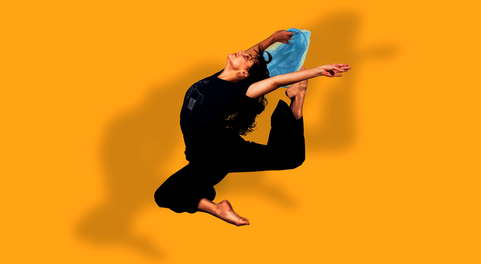 Tänzerin springt in einer ausdrucksstarken Pose vor gelbem Hintergrund, ein Bein angewinkelt, Arme weit ausgestreckt, hält ein blaues Tuch.