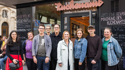 Gruppe von neun Personen vor einem Café mit Schriftzug "Kuchenamt" auf der Glasfront