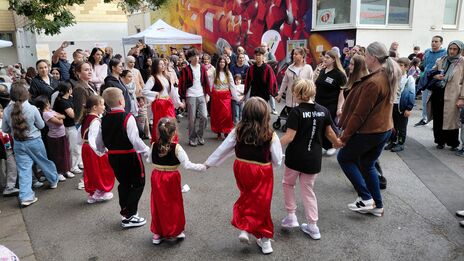 Kinder und Erwachsene halten sich im Kreis an den Händen und tanzen auf einem Hof vor einem Haus mit bunter Graffiti-Wand mit einer Menschenmenge im Hintergrund