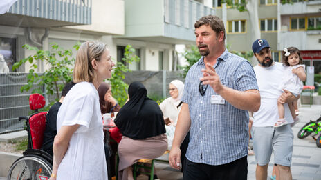 Ein wohnpartner-Mitarbeiter spricht mit einer Frau in eineFrauen m weißen Kleid im Innenhof einer Wohnhausanlage. Im Hintergrund sitzen mehrere Menschen an einem Tisch, darunter drei Frauen mit Kopftuch und ein Junge im Rollstuhl, ein Mann hält ein Kind auf dem Arm.