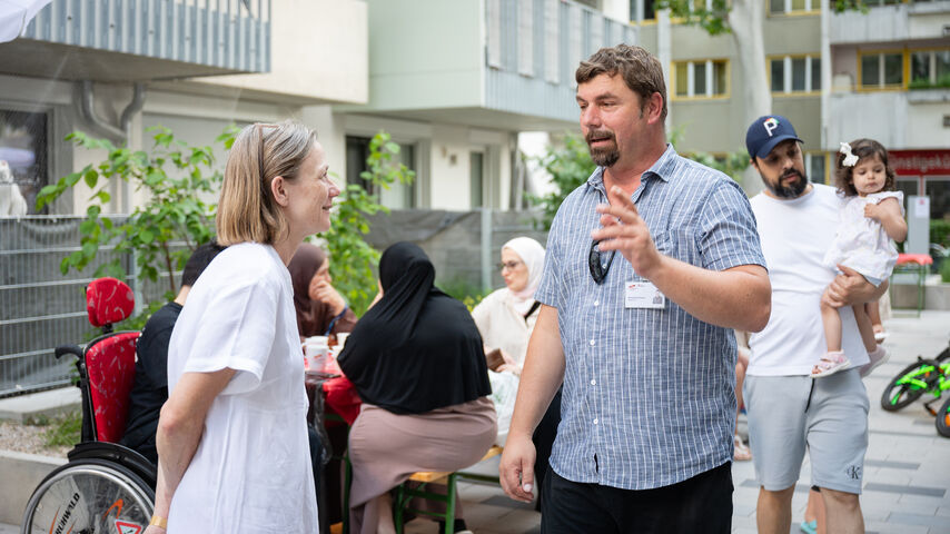 Ein wohnpartner-Mitarbeiter spricht mit einer Frau in eineFrauen m weißen Kleid im Innenhof einer Wohnhausanlage. Im Hintergrund sitzen mehrere Menschen an einem Tisch, darunter drei Frauen mit Kopftuch und ein Junge im Rollstuhl, ein Mann hält ein Kind auf dem Arm.