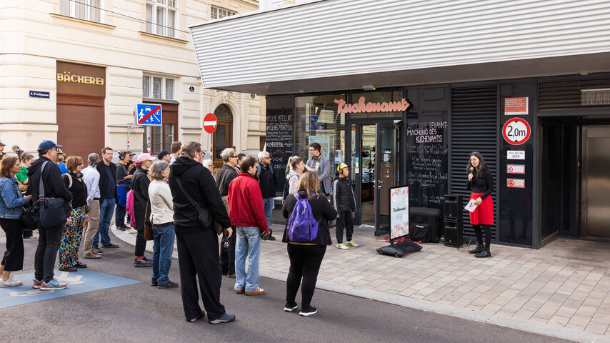Gruppe von Menschen steht auf einem Bürgersteig vor einem modernen Gebäude mit Schaufenster und hört einer Frau zu, die vor dem Eingang spricht