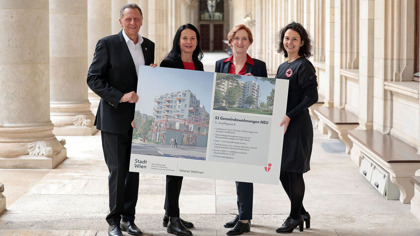 Drei weibliche und eine männliche Person stehen in den Arkaden des Wiener Rathauses und halten gemeinsam ein Schild auf dem Visualisierungen des Gemeindebau NEU zu sehen sind, sowie ein wenig Text