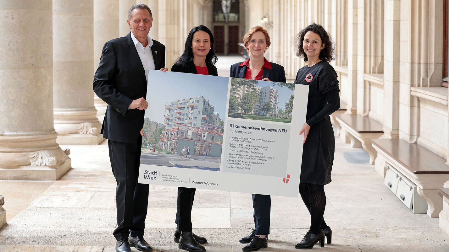 Drei weibliche und eine männliche Person stehen in den Arkaden des Wiener Rathauses und halten gemeinsam ein Schild auf dem Visualisierungen des Gemeindebau NEU zu sehen sind, sowie ein wenig Text