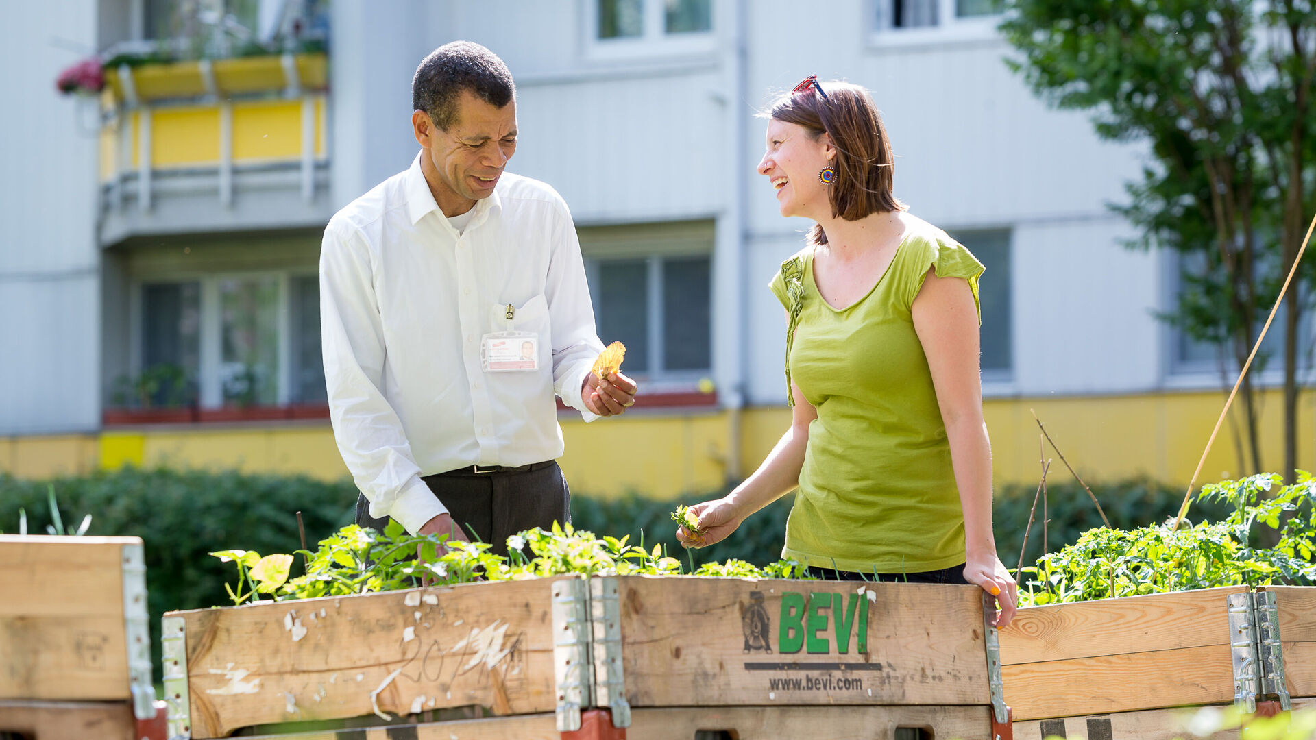 Ein wohnpartner-Mitarbeiter und eine Frau stehen vor einem mobilen Beet in einem Innenhof. Der Mann hält ein Blatt in der Hand, die Frau lacht.