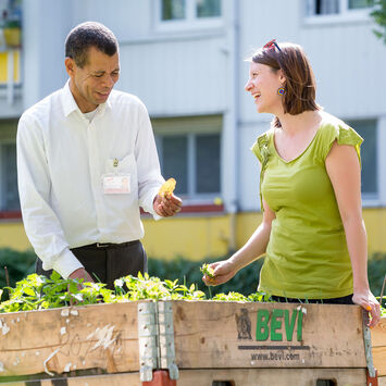 Ein wohnpartner-Mitarbeiter und eine Frau stehen vor einem mobilen Beet in einem Innenhof. Der Mann hält ein Blatt in der Hand, die Frau lacht.