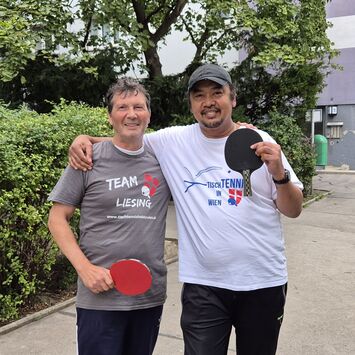 Zwei Männer stehen im Freien neben einem Tischtennis-Tisch, haben Tischtennis-Shirts an und halten Tischtennis-Schläger in die Höhe.