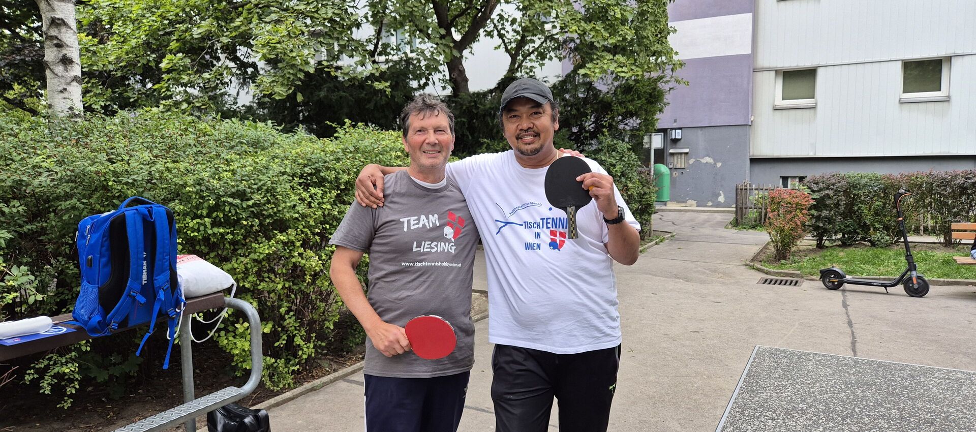Zwei Männer stehen im Freien neben einem Tischtennis-Tisch, haben Tischtennis-Shirts an und halten Tischtennis-Schläger in die Höhe.