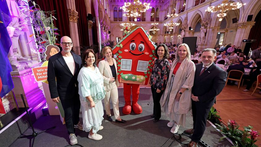 Gruppe von neun Personen steht in festlich beleuchtetem Saal neben wienwohntbesser-Maskottchen, ein rotes Haus mit Gesicht und Blumenschmuck, sie halten ein Schild mit Text 'Wir sorgen für ein gutes Miteinander'