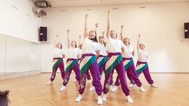 Gruppe von Jugendlichen tanzt synchron in einem hellen Proberaum, Arme nach oben gestreckt.