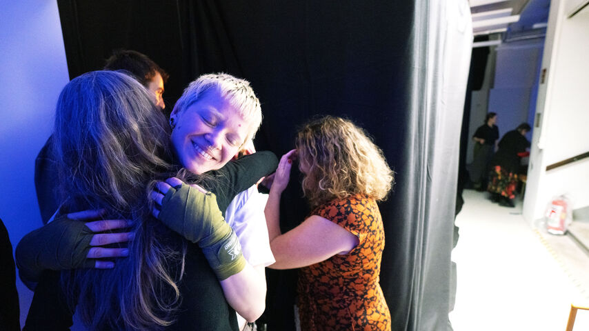 Drei Personen umarmen sich vor einem schwarzen Vorhang in einem hell beleuchteten Raum nach einer Theaterpremiere